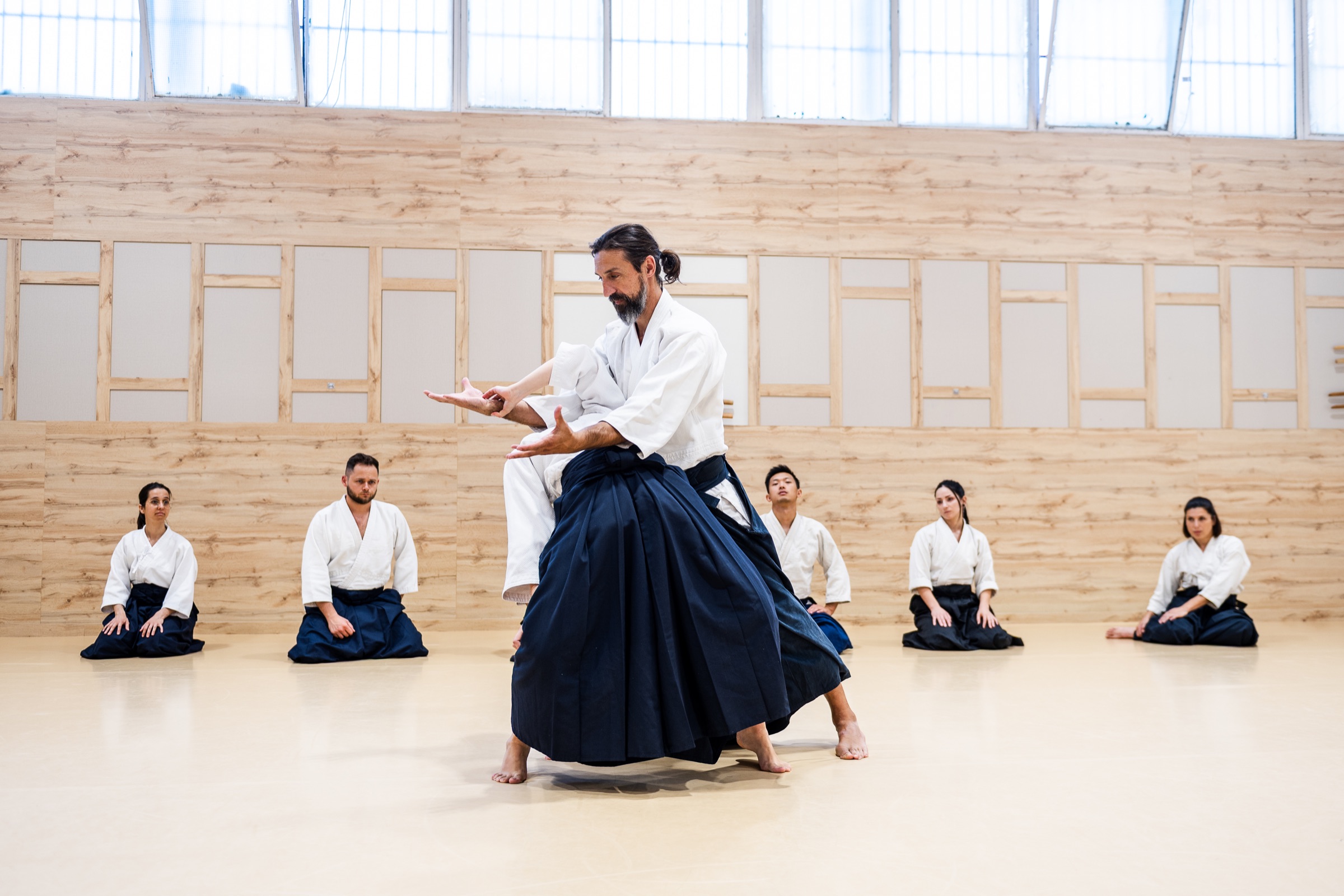 The instructor shows a technique to the class, seated around the mat.